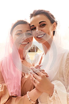 Close up portrait of happy charming bride in a white dress with a white veil and a bridesmaid in a beige dress with pink