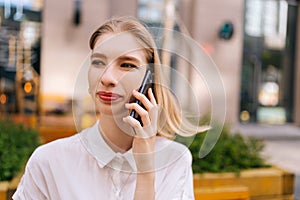 Close-up portrait of happy charming young blonde woman talking on smartphone sitting on bench on city street, smiling