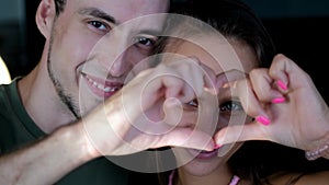 Close up portrait happy couple faces looking through joined fingers making heart shape. St Valentines Day celebration