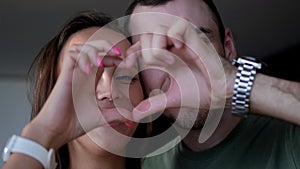 Close up portrait happy couple faces looking through joined fingers making heart shape. St Valentines Day celebration