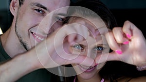 Close up portrait happy couple faces looking through joined fingers making heart shape. St Valentines Day celebration