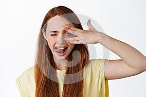 Close up portrait of happy cute girl with long red hiar, winking and showing tongue, open eye peek through fingers and
