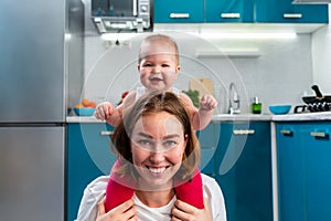 Close-up portrait of a happy mother and her baby sitting on her shoulders. The kitchen is in the background. The concept of family