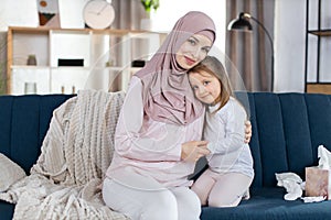 Close up portrait of happy Muslim family, mother and little daughter, sitting together on blue sofa and hugging, posing