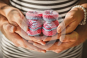 Close up portrait of happy pregnant woman together with husband holding small baby shoes, hugging in summer park