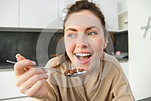 Close up portrait of happy smiling brunette in bathrobe, holding cereals in spoon, eating breakfast with milk and