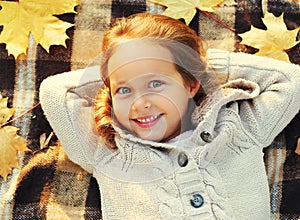 Close up portrait of happy smiling little girl child lying on a plaid with yellow maple leaves in autumn day