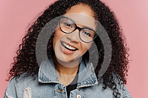 Close up portrait of happy smiling woman with dark curly Afro hairstyle, tilts head, wears optical glasses and denim jacket,