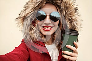 Close up portrait of happy smiling woman stretching hand for taking selfie with coffee cup wearing red jacket