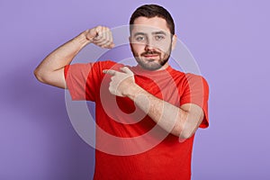 Close up portrait of happy strong man dresses casual red t shirt showing his biceps on arm and pointing with index finger, male