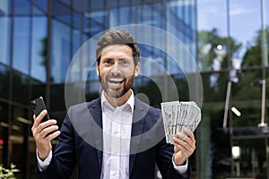 Close-up portrait of a happy, successful and rich young businessman standing outside an office building, holding a phone