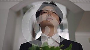 Close-up portrait of happy young Asian groom standing with bouquet indoors looking away. Confident serious man holding