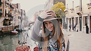 Close-up portrait of happy young beautiful European woman tourist in hat smiling standing by famous Venice canal, Italy.