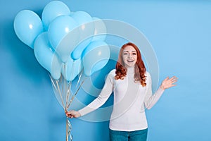 Close up portrait happy young beautiful European woman in white shirt and jeans, looks surprised with blue party balloon, keeps