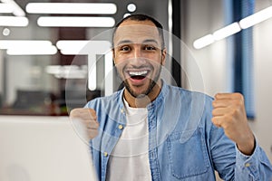 Close-up portrait of a happy young hispanic man sitting in a modern office in front of a laptop and looking at the