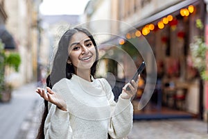 Close-up portrait of a happy young Indian woman standing on a city street, holding a phone in her hands, happy with the