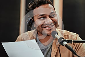 Close up portrait of happy young male radio host in headphones smiling aside while talking, broadcasting in studio