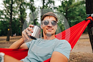 Close-up portrait of a happy young man in sunglasses lying in a hammock with a cup of hot drink in his hand, looking away and