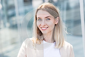 Close-up portrait of a happy young woman standing outdoors and smiling on blurred urban background looking at camera. Female