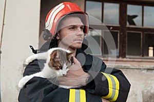 Close-up portrait of heroic fireman in protective suit and red helmet holds saved cat in his arms. Firefighter in fire