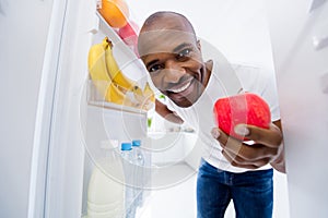 Close-up portrait of his he nice attractive cheerful cheery guy looking in fridge taking red ripe apple snack health