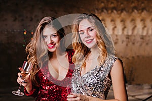 Close-up portrait of interested good-looking ladies standing on dark background with wine. Adorable female models in