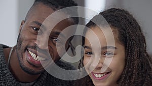 Close-up portrait of laughing joyful African American teen daughter and father posing indoors at home on weekend