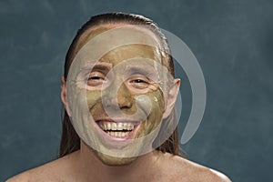 Close-up portrait of laughing pleasured man with wet long hair in green clay mask on his face waiting of spa procedures