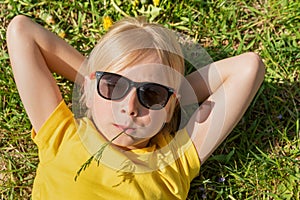 Close up portrait of little blond boy wearing yellow t-shirt and sunglasses lies on grass. Child rests on dandelions filed with