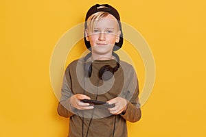 Close up portrait of little child boy plays with his phone,keeps headhones around neck, dresses green shirt and backwards hat,