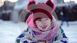 Close up Portrait Little Child Pretty Girl Kid Smiling Looking at Camera play red hat lie on Stomack. Snow Park Outdoor