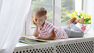 Close up portrait of little cute girl reading book on windowsill at home.