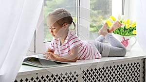 Close up portrait of little cute girl reading book on windowsill at home.