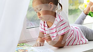 Close up portrait of little cute girl reading book on windowsill at home.