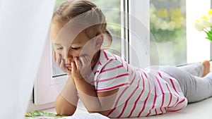 Close up portrait of little cute girl reading book on windowsill at home.