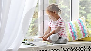 Close up portrait of little cute girl reading book on windowsill at home.