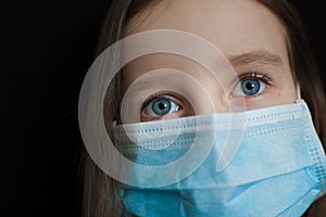 Close-up portrait of little defenseless girl on black backgound in blue disposable mask for protection of Coronavirus