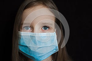 Close-up portrait of little defenseless girl on black backgound in blue disposable mask for protection of Coronavirus