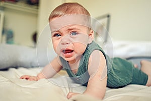 Close up portrait of little funny cute blonde infant boy child toddler with blue eyes in green linen bodysuit crying