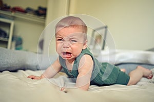 Close up portrait of little funny cute blonde infant boy child toddler with blue eyes in green linen bodysuit crying