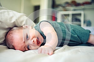 Close up portrait of little funny cute blonde infant boy child toddler with blue eyes in green linen bodysuit crying