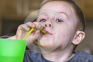 Close up portrait of little kid in the kitchen at home. Pretty child drinks a cold drink from a green plastic cup through a straw.