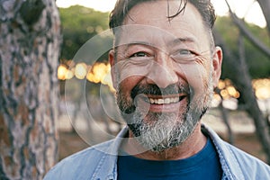 Close up portrait looking on camera of adult mature man with beard and big smile at the park. Aged male person in cheerful