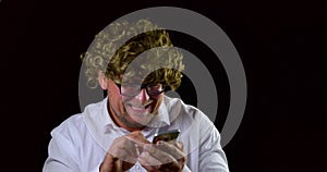 Close-up portrait of a mad man with glasses and curly hair using a mobile phone for the Internet on a black background.