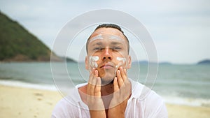 Close-up portrait man apply sun cream protection lotion, looking at camera. Funny man on beach near sea smearing