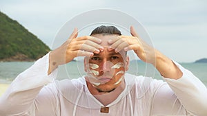 Close-up portrait man apply sun cream protection lotion, looking at camera.Stylish men on beach near sea smearing