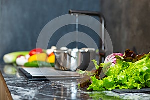 Close-up portrait of a man in a brown apron pouring water into a pot in the kitchen. Washed vegetables lie on the sink.