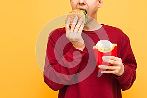 Close-up portrait of a man eating bad food, biting a burger and holding french fries in his hands on a yellow background. Guy with