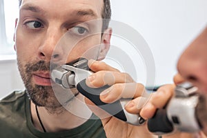 Close up portrait of a man shaving beard or stubble with an electric razor. Reflection in mirror of an unshaven face of guy with