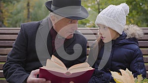 Close-up portrait of a mature Caucasian man in classic clothes sitting on the bench with his granddaughter and reading a